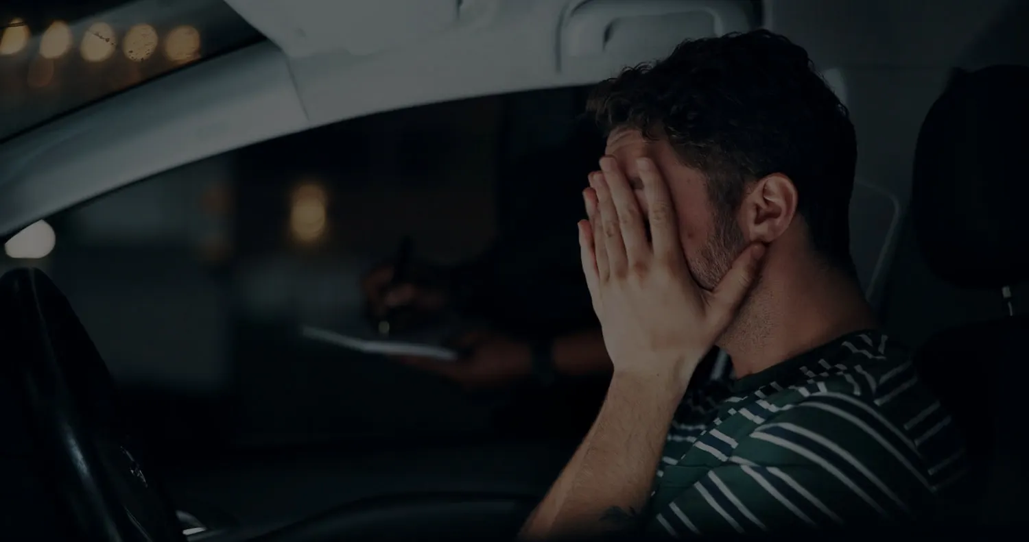 Driver covering his face in his hands while a police officer writes notes outside the car at night